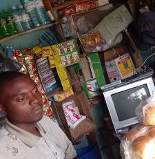 The young man, Martin Kakuru, in his store with a computer screen that he has repaired, and a pile of bread rolls he has made. 