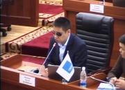 MP Dastan Bekeshev at a desk, addressing the meeting, through a microphone