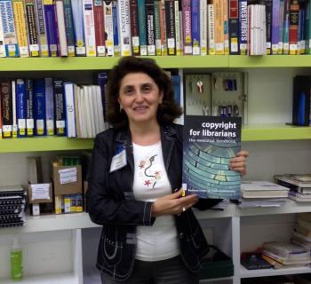 Full frontal photo of Hasmik Galystan, EIFL Copyright Coordinator in Armenia holding EIFL publication 'Copyright for Librarians'. Hasmik is standing in front of library shelves where she works at the American University of Armenia. 