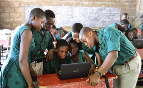 Children in a school classroom crowded around a laptop computer. 